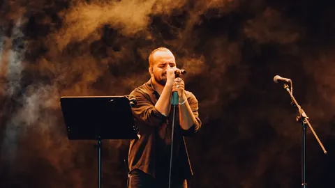 Faheem Abdullah singing into a microphone on a smoky stage lit in orange, with a music stand beside him