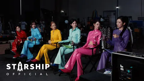 IVE's six members on director's chairs in a studio, each in a shiny suit of a different colour