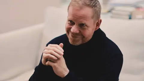 Max Richter smiling in a black turtleneck, hands clasped, seated on a pale sofa in a softly lit room