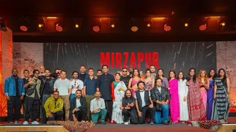Mirzapur: The Movie cast and crew lined up on stage under orange lights in front of the red Mirzapur title