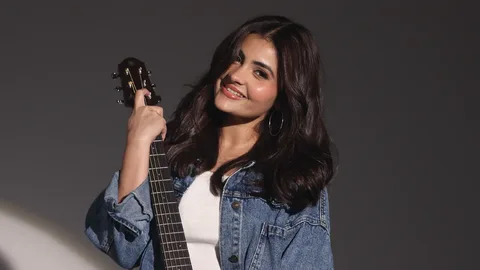 Noor Chahal smiling in a denim jacket and white top, holding an acoustic guitar by its neck against a grey wall