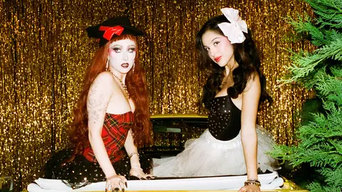 Chappell Roan in red tartan and Olivia Rodrigo with a white bow, seated on a car before a gold tinsel curtain