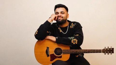 Parth Srivastava smiling in a black floral sweater, leaning on an acoustic guitar against a plain cream wall